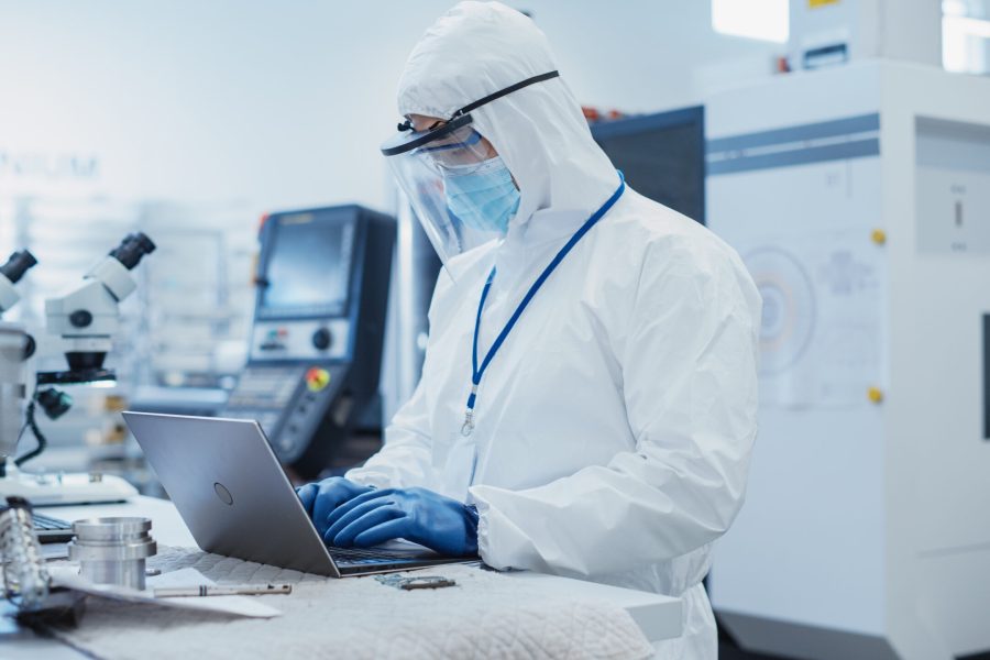 Electronic Manufacturing Factory: Engineer in Sterile Coverall Working on Laptop Computer, Examining a Circuit Board with Microchips and Testing New Electrical Equipment.
