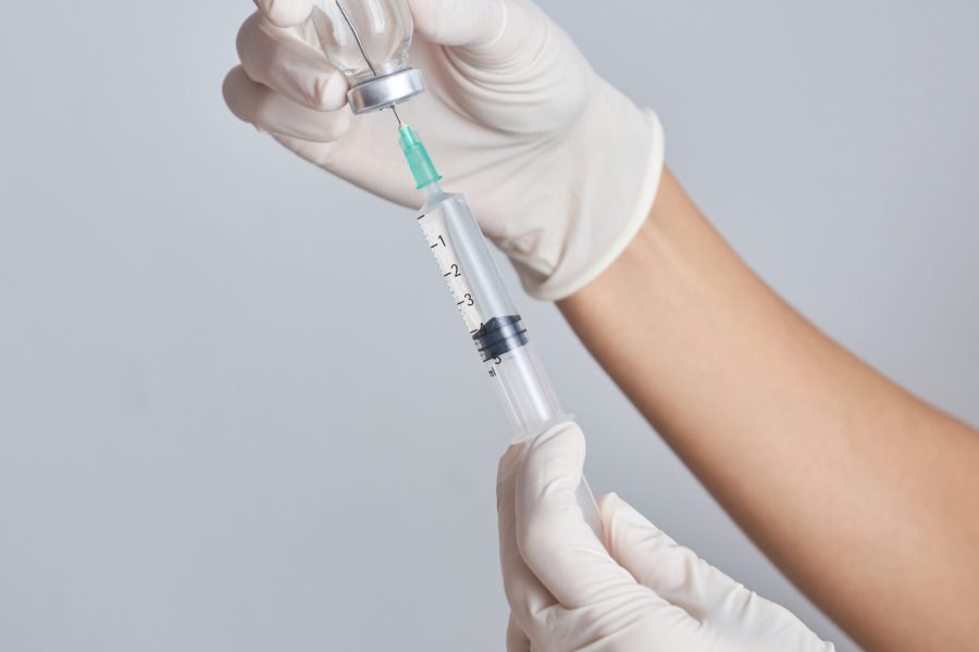 Hands of crop unrecognizable doctor in protective gloves drawing up vaccine from vial into syringe against white background in room