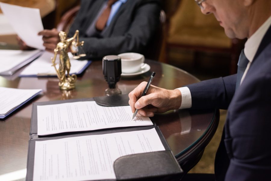 Mature businessman or jurist signing document by workplace Mature businessman or jurist in formalwear signing juridical document by workplace against his colleague looking through papers